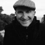Black and white portrait of a smiling senior man wearing a flat cap outdoors.