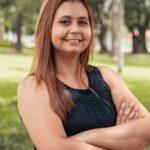Confident woman with arms crossed standing in a lush green park on a sunny day.
